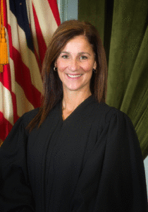 Hon. Paula Roscioli is pictured in a black formal robe in front of the American flag