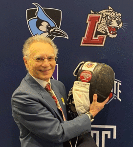 Mayer is wearing a blue jacket and maroon tie. He is holding a Lafayette item in front of a backdrop with the Lafayette logo and other college logos