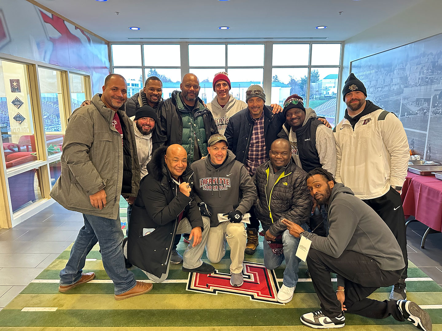 Lafayette College Football alumni pose with Coach John Troxell '94 in Kirby Football House. Coach Troxell is wearing a Lafayette Football sweatshirt. They are smiling at the camera. 